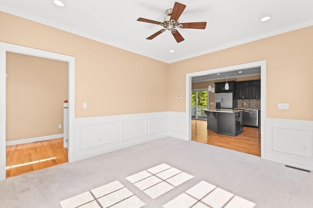 unfurnished living room featuring light carpet, ornamental molding, a wainscoted wall, a ceiling fan, and recessed lighting