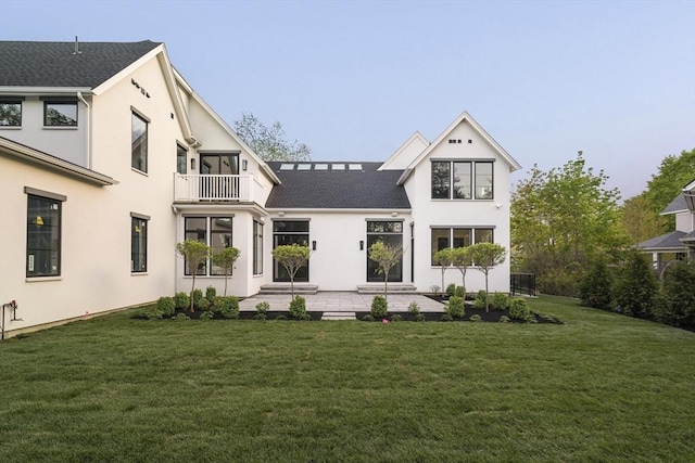 back of house with a balcony, a lawn, a patio area, stucco siding, and roof with shingles