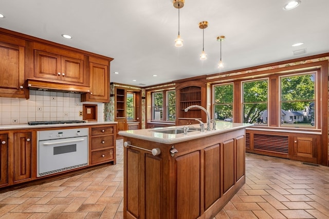 kitchen with white oven, pendant lighting, a kitchen island with sink, sink, and decorative backsplash