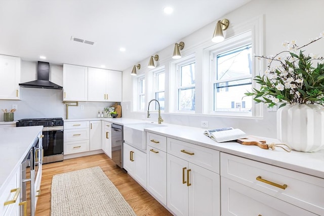 kitchen with wall chimney exhaust hood, sink, light wood-type flooring, stainless steel appliances, and white cabinets