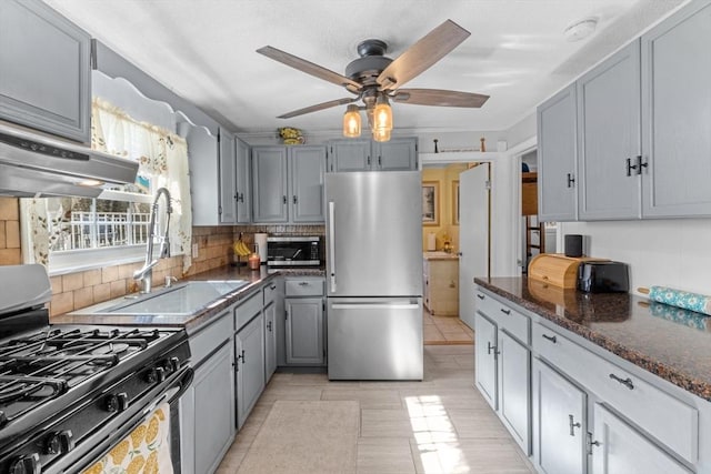 kitchen with gray cabinets, a sink, under cabinet range hood, gas range oven, and freestanding refrigerator