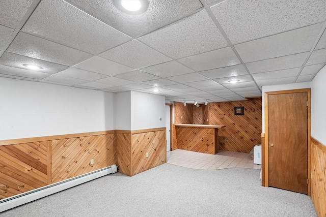 basement featuring carpet, a drop ceiling, baseboard heating, and wood walls