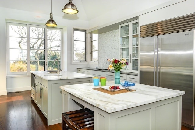 kitchen featuring light stone counters, sink, built in refrigerator, a kitchen island with sink, and decorative light fixtures