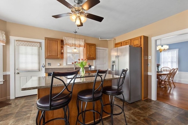 kitchen with brown cabinets, stainless steel fridge with ice dispenser, a breakfast bar area, ceiling fan with notable chandelier, and a kitchen island
