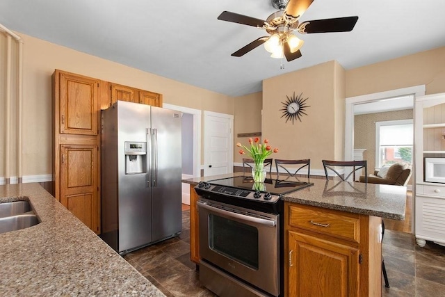 kitchen featuring ceiling fan, appliances with stainless steel finishes, light stone countertops, brown cabinetry, and a kitchen island