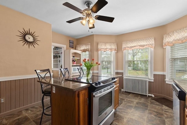 kitchen with stone finish floor, a ceiling fan, appliances with stainless steel finishes, wainscoting, and radiator heating unit