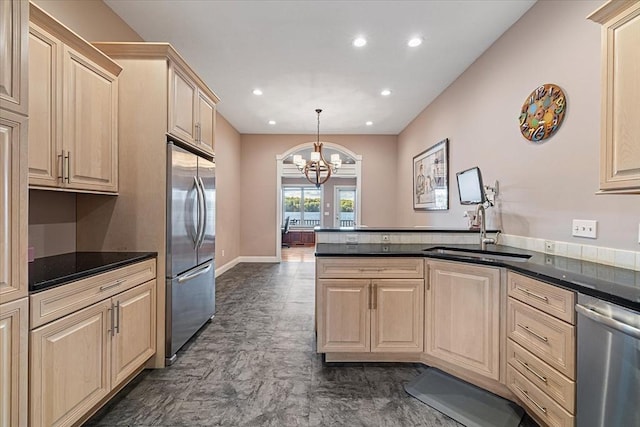 kitchen featuring light brown cabinetry, dark countertops, a sink, an inviting chandelier, and stainless steel appliances