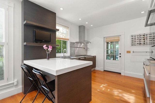 kitchen featuring wall chimney range hood, a sink, light wood-type flooring, stainless steel gas stovetop, and recessed lighting