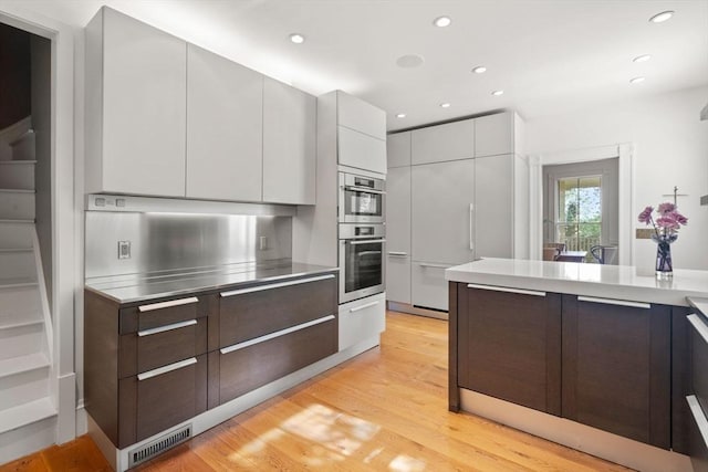 kitchen featuring stainless steel countertops, modern cabinets, double oven, light wood-style floors, and dark brown cabinets