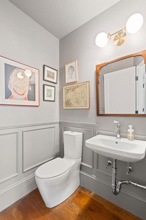 half bath featuring wood finished floors, toilet, a decorative wall, a sink, and wainscoting