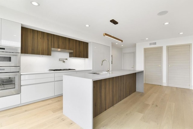 kitchen with white cabinets, sink, hanging light fixtures, an island with sink, and stainless steel double oven