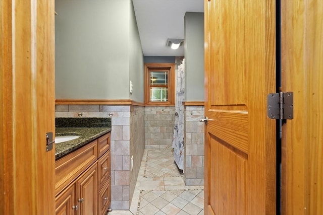 full bathroom with tile walls, tile patterned flooring, vanity, a wainscoted wall, and tiled shower