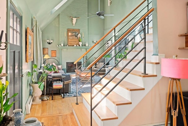 staircase featuring wood-type flooring, high vaulted ceiling, a ceiling fan, a brick fireplace, and baseboard heating