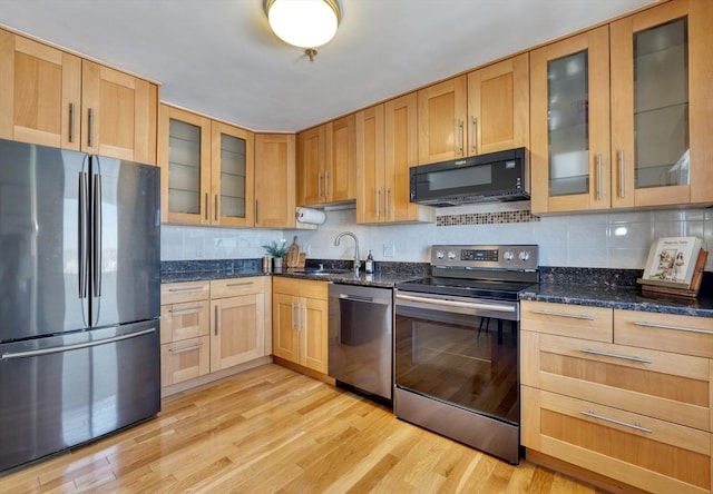 kitchen featuring a sink, light wood-style floors, appliances with stainless steel finishes, dark stone counters, and glass insert cabinets