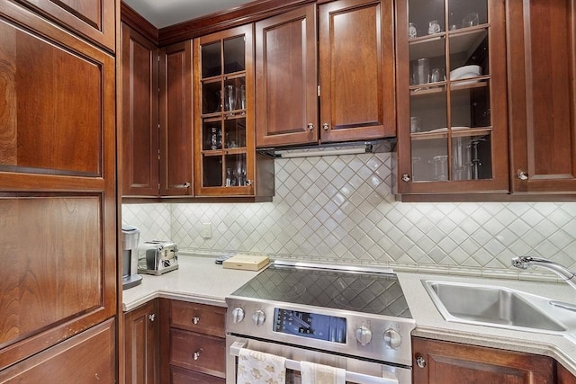 kitchen with glass insert cabinets, light countertops, stainless steel electric stove, tasteful backsplash, and a sink