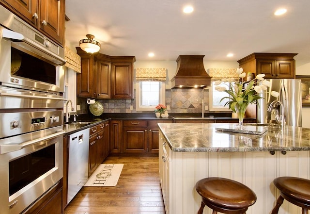 kitchen with premium range hood, a kitchen bar, a center island with sink, stainless steel appliances, and wood-type flooring