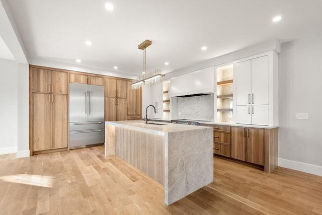 kitchen with decorative light fixtures, white cabinetry, an island with sink, custom range hood, and built in refrigerator