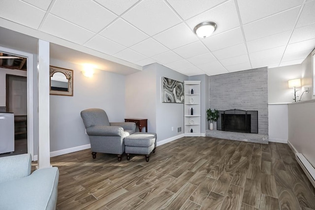 sitting room featuring baseboards, wood finished floors, baseboard heating, a paneled ceiling, and a brick fireplace