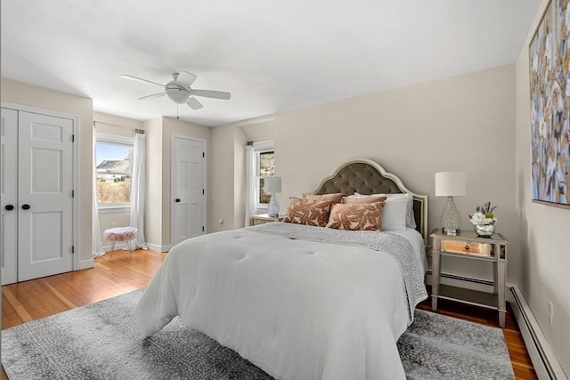 bedroom featuring baseboard heating, two closets, a ceiling fan, baseboards, and wood finished floors