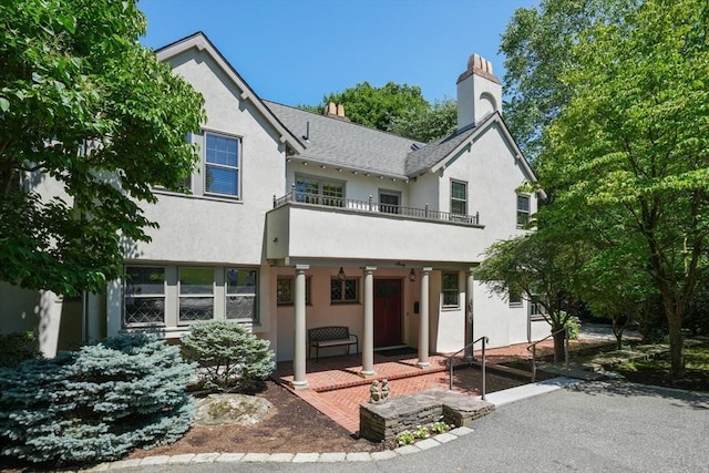 view of front facade featuring a patio, a chimney, stucco siding, a shingled roof, and a balcony