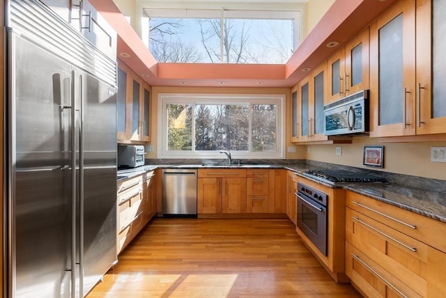 kitchen with dark stone countertops, brown cabinetry, glass insert cabinets, appliances with stainless steel finishes, and light wood-type flooring