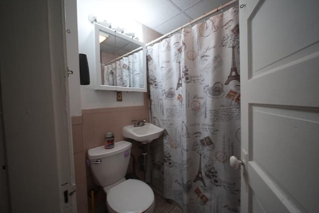 full bath with a sink, a drop ceiling, toilet, a wainscoted wall, and tile walls