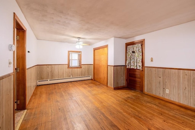 empty room featuring a textured ceiling, light wood-type flooring, baseboard heating, wooden walls, and ceiling fan