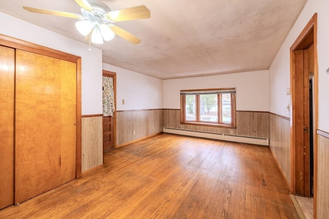 unfurnished bedroom with a baseboard heating unit, light hardwood / wood-style floors, ceiling fan, and wood walls