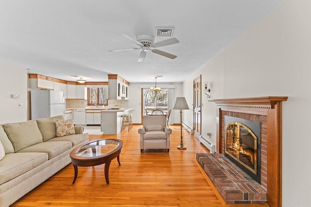 living area featuring visible vents, baseboard heating, light wood-type flooring, a fireplace, and ceiling fan with notable chandelier