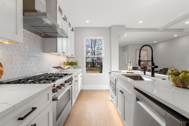 kitchen with white cabinetry, a sink, light wood-style floors, appliances with stainless steel finishes, and wall chimney exhaust hood