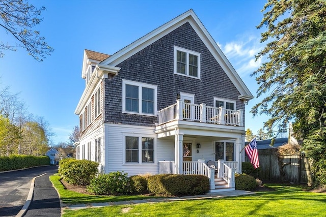 shingle-style home featuring covered porch, fence, aphalt driveway, a front lawn, and a balcony