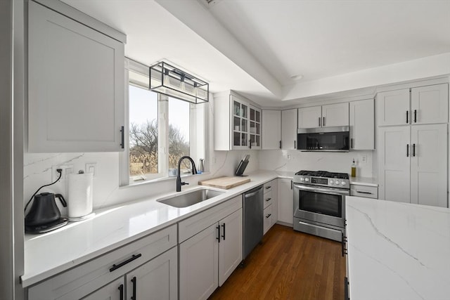 kitchen with a sink, dark wood finished floors, appliances with stainless steel finishes, glass insert cabinets, and light stone countertops