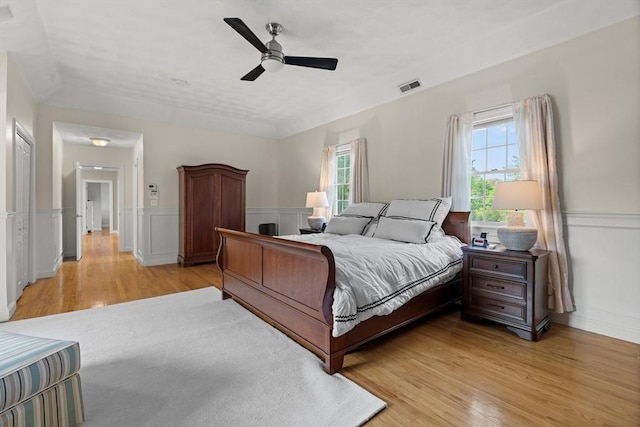 bedroom with multiple windows, light wood-type flooring, a wainscoted wall, and ceiling fan