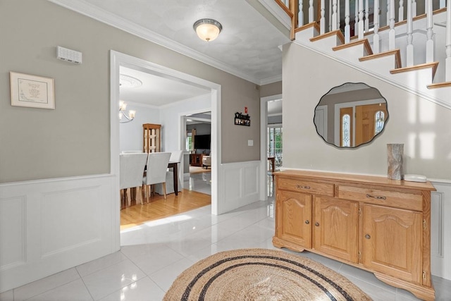 entryway with light tile patterned flooring, ornamental molding, a chandelier, wainscoting, and a decorative wall