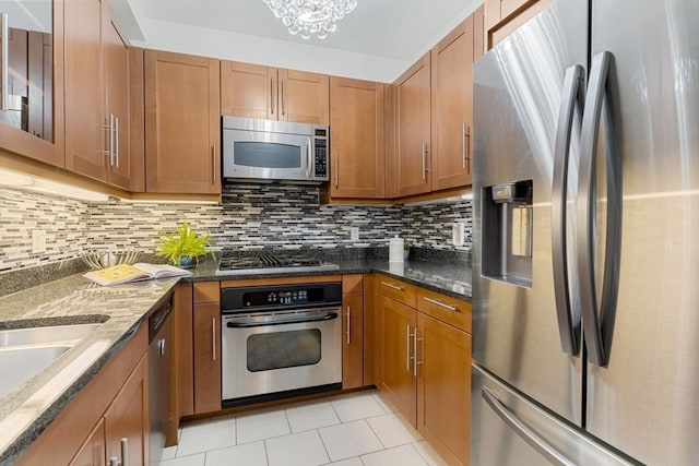 kitchen with light tile patterned flooring, stainless steel appliances, decorative backsplash, an inviting chandelier, and dark stone countertops