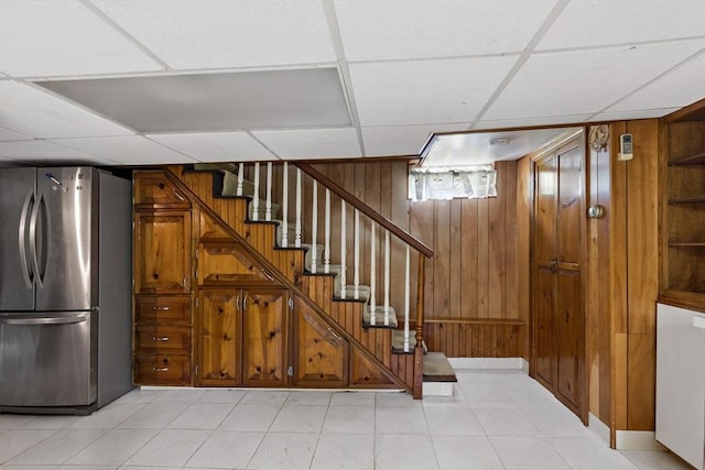 basement with freestanding refrigerator, a drop ceiling, wood walls, and stairs