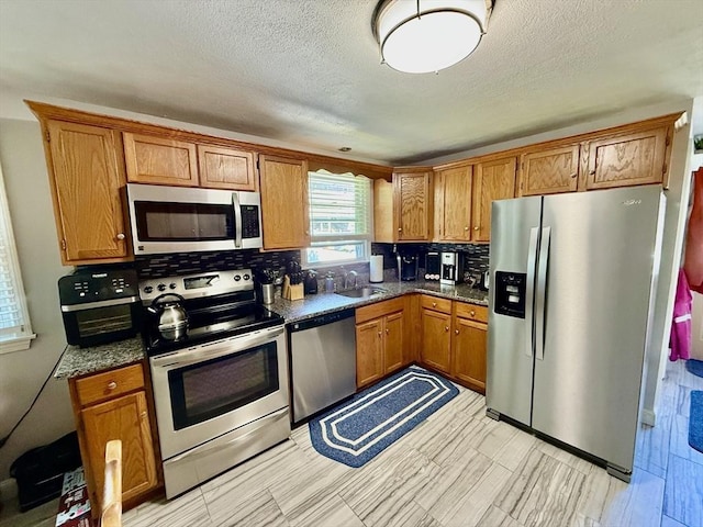 kitchen with appliances with stainless steel finishes, a sink, brown cabinetry, tasteful backsplash, and dark countertops