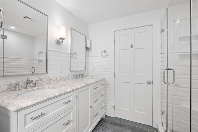 full bath featuring visible vents, double vanity, a stall shower, tile walls, and a sink