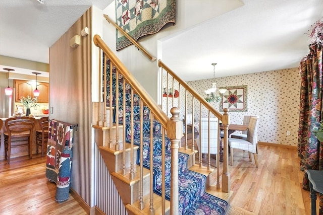 stairway featuring wallpapered walls, wood finished floors, baseboards, and a chandelier