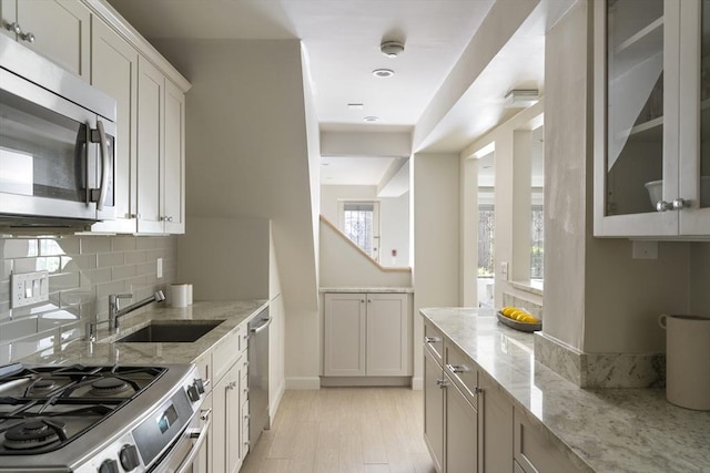 kitchen with appliances with stainless steel finishes, a sink, light stone counters, decorative backsplash, and glass insert cabinets