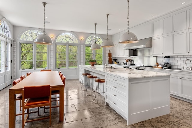 kitchen with hanging light fixtures, wall chimney exhaust hood, a kitchen island with sink, and backsplash