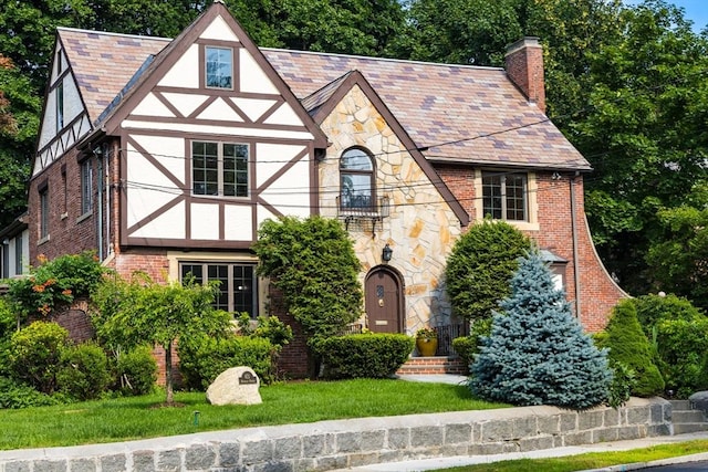 tudor home featuring brick siding, stucco siding, a front lawn, a chimney, and a high end roof