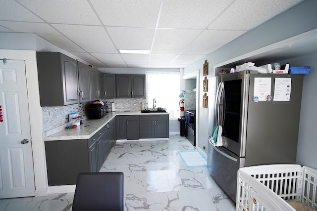 kitchen with gray cabinets, a paneled ceiling, stainless steel refrigerator, sink, and backsplash