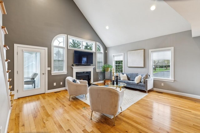 living area with a warm lit fireplace, high vaulted ceiling, baseboards, and light wood-style flooring