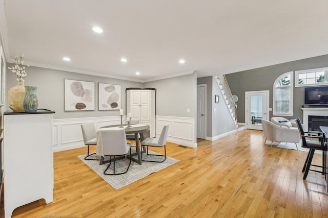 dining space featuring ornamental molding, a glass covered fireplace, light wood-style floors, stairway, and a wainscoted wall