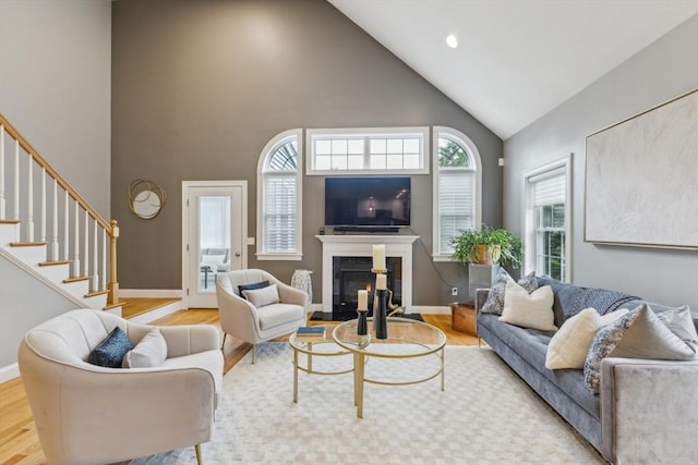 living area with stairs, a glass covered fireplace, wood finished floors, baseboards, and high vaulted ceiling