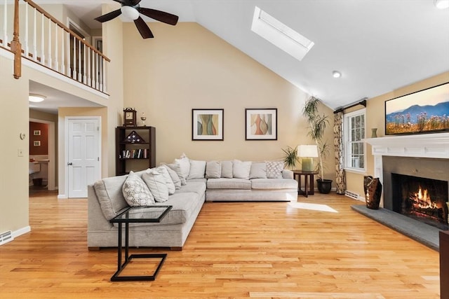 living area with ceiling fan, a skylight, light wood-type flooring, high vaulted ceiling, and a warm lit fireplace