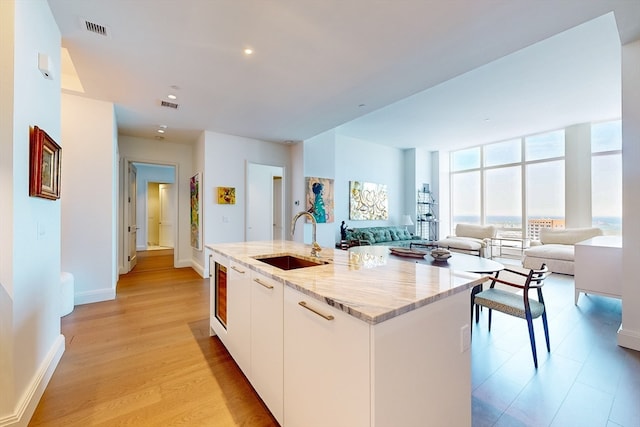 kitchen with light hardwood / wood-style flooring, a kitchen island with sink, sink, light stone counters, and white cabinets