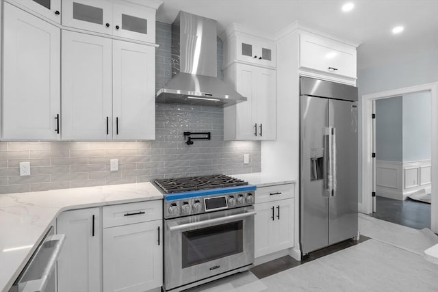 kitchen with light stone countertops, backsplash, high end appliances, wall chimney range hood, and white cabinets