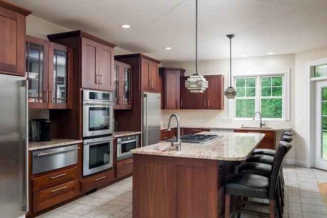 kitchen with sink, stainless steel appliances, light stone counters, pendant lighting, and a kitchen island with sink